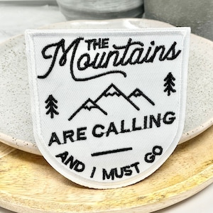 May include: White embroidered patch with black text that reads "The Mountains Are Calling And I Must Go". The patch features a mountain range and pine trees.