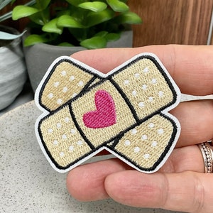 Peut inclure: Un patch brodé en forme de deux bandages se chevauchant avec un cœur rose au centre.