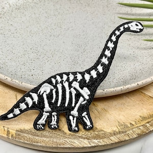 Könnte beinhalten: Schwarzer und weißer gestickter Dinosaurierskelett-Patch. Der Patch befindet sich auf einem weißen Teller mit einer gesprenkelten Oberfläche und einer Holzoberfläche.