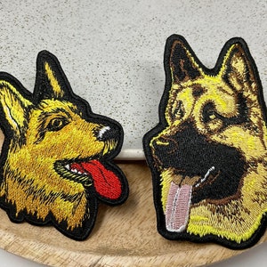 Könnte beinhalten: Zwei gestickte Aufnäher mit Schäferhundköpfen. Einer zeigt den Hund mit herausgestreckter Zunge, der andere im Profil. Beide Aufnäher haben schwarze Ränder und liegen auf einem Holztablett.