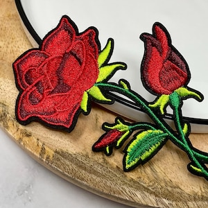 May include: Embroidered red rose patches with green leaves and stems. One rose is fully bloomed, the other is a bud. The patches are on a wooden surface next to a white plate with a black rim. These patches are ideal for clothing or craft projects.