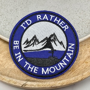 Peut inclure: Un écusson brodé bleu et blanc avec le texte "I'd Rather Be In The Mountain". L'écusson représente une chaîne de montagnes blanches avec une silhouette noire d'un arbre et un poteau indicateur.