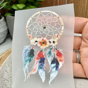 Könnte beinhalten: Ein weißer Traumfänger mit goldenen Perlen und Federn. Der Traumfänger hat ein florales Design mit rosa, roten und gelben Blumen. Die Federn sind blau, grün und rot.