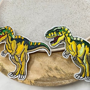 Peut inclure: Deux patchs brodés thermocollants représentant des dinosaures verts et jaunes. Les dinosaures sont tournés dans des directions différentes et ont des dents blanches.