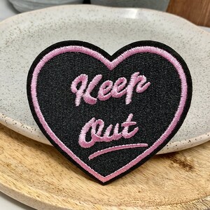 Peut inclure: Un patch en forme de cœur noir avec des coutures roses. Le texte "Keep Out" est écrit en rose sur le patch.