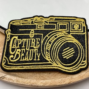 Peut inclure: Un patch brodé noir et or représentant un appareil photo vintage avec le texte "Capture Beauty".