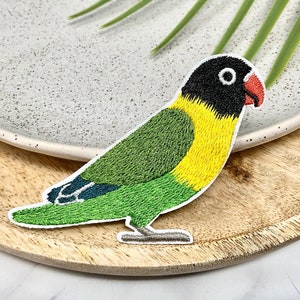 Peut inclure: Écusson brodé d'un oiseau coloré, avec une tête noire, un bec rouge, une poitrine jaune et des ailes et une queue vertes. Le patch est sur fond blanc avec un plateau en bois et une feuille verte.