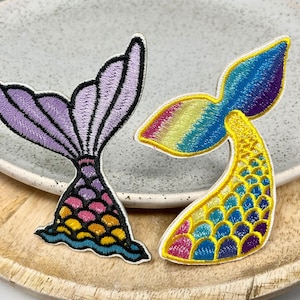 Könnte beinhalten: Zwei gestickte Meerjungfrauen-Schwanz-Patches. Einer ist lila mit einem regenbogenfarbenen Schwanz und der andere ist gelb mit einem blauen und violetten Schwanz.