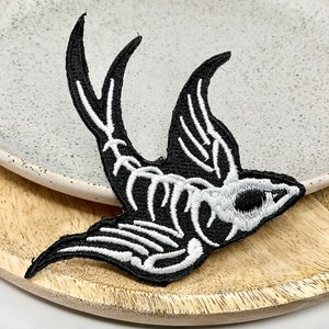 May include: Black and white embroidered iron-on patch of a skeleton bird with its wings spread. The bird is in a flying position.