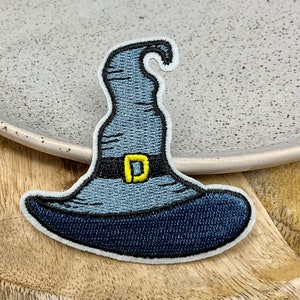 Peut inclure: Écusson brodé en forme de chapeau de sorcière bleu, noir et jaune. Le chapeau a une couronne pointue, un large bord et une bande noire avec une boucle jaune. L'écusson a une bordure blanche.