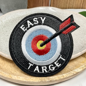 Peut inclure: Un patch brodé noir et blanc avec une cible rouge et blanche et une flèche passant par le centre. Le texte "EASY TARGET" est écrit autour de la cible.
