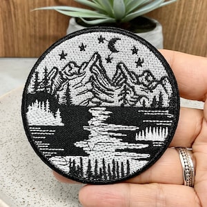May include: Black and white embroidered patch featuring a mountain range, lake, and moon with stars. The patch is circular and has a black background with white embroidery.