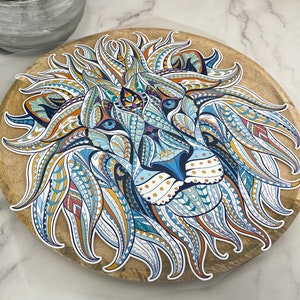 Op de afbeelding: Een kleurrijk, ingewikkeld mandala-achtig ontwerp van een leeuwenkop op een houten onderzetter. De leeuwenmanen is gemaakt van draaiende patronen in tinten blauw, oranje en wit.