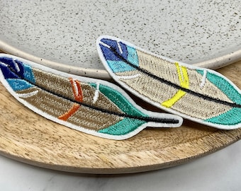Puede incluir: Dos parches bordados con forma de pluma con borde blanco. Las plumas tienen una base marrón con detalles turquesa, azul, naranja y amarillo. Los parches están sobre una bandeja de madera y un plato de cerámica moteado.