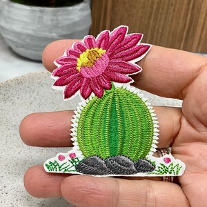 May include: Embroidered iron-on patch featuring a pink flower blooming on top of a green cactus with grey rocks. The cactus is surrounded by green grass and small pink flowers.