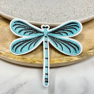 May include: Embroidered dragonfly patch with turquoise and black wing details. The dragonfly has a white border and is set against a wooden surface. This decorative patch can be ironed on or sewn onto clothing or accessories.