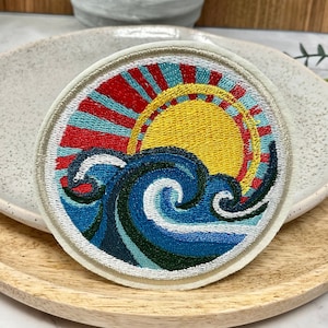 Peut inclure: Un patch rond brodé avec un motif coloré d'un soleil couchant sur une vague bleue. Le soleil est jaune avec des rayons rouges et bleus. La vague est bleue avec de l'écume blanche.