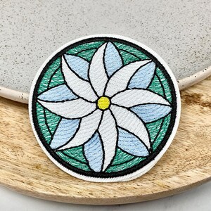 Peut inclure: Patch brodé représentant une fleur blanche avec un centre jaune, entourée d'un motif circulaire vert et bleu.