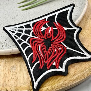 May include: Embroidered patch with a red spider on a black background, framed by a white spiderweb design. The patch has an unusual, angular shape, suitable for clothing or accessories.