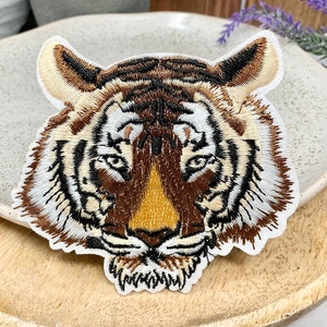 May include: Embroidered tiger face patch with brown, white, and black stripes. The tiger's eyes are open and the mouth is closed. The patch is on a light brown wooden surface.