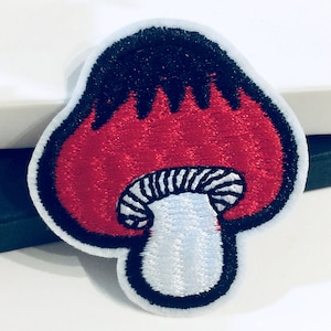 Pode incluir: Patch de cogumelo bordado em vermelho e branco com contorno preto. O cogumelo tem um caule branco com listras pretas e um chapéu vermelho com chamas pretas.