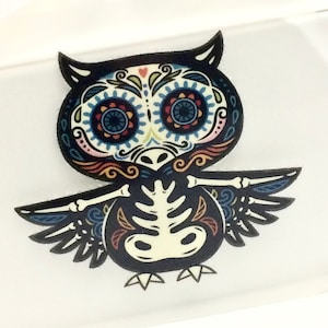 May include: A black and white owl with colorful sugar skull details. The owl has large, round eyes with blue, red, and yellow accents. The owl's wings are spread out, and its body is decorated with intricate floral patterns.