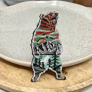May include: An embroidered iron-on patch depicting a brown bear with a mountain scene within its body. The bear is standing on a green background with trees and a lake. The bear's fur is orange and brown, and the mountains are grey and white.