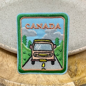 May include: A blue and green embroidered patch with a cartoon image of a vintage camper van driving on a road with trees and clouds. The text "CANADA" is written in orange letters on the patch.