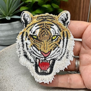 May include: Close-up of an embroidered tiger face patch with orange, black, and white stripes. The tiger has a fierce expression with a wide open mouth and sharp teeth.