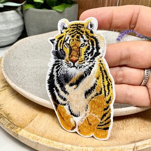 May include: Embroidered tiger patch with a detailed design in orange, black, and white. The patch shows a tiger with a fierce expression, ideal for jackets, bags, or other fabric items.