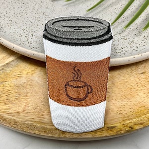 May include: Embroidered iron-on patch of a white coffee cup with a brown coffee design and steam coming from the top. The coffee cup is on a brown wooden surface.