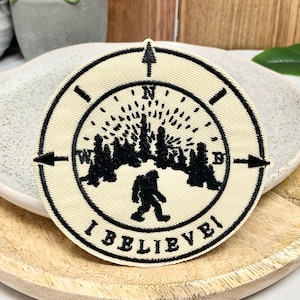 May include: A beige embroidered patch with a black compass design. The compass points north and features a silhouette of a Bigfoot walking through a forest. The text "I Believe" is written around the compass.