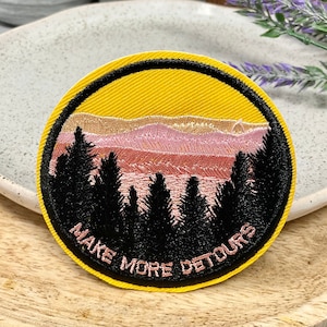 Peut inclure: Un patch brodé jaune, rose et noir avec une scène de coucher de soleil et le texte "Make More Detours".