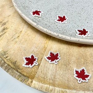 Può includere: Un set di toppe da stirare a forma di foglia d'acero rossa su una superficie di legno e un piatto bianco.