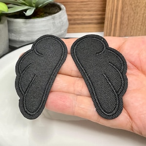 Peut inclure: Deux écussons d'ailes en tissu noir avec des détails cousus. Les écussons ont la forme d'ailes stylisées, avec des bords incurvés et des lignes internes pour représenter les plumes. Les ailes sont en tissu texturé, probablement pour l'artisanat ou l'embellissement.