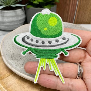 Peut inclure: Patch brodé vert et blanc représentant un OVNI de dessin animé avec une fenêtre jaune. L'OVNI a un faisceau de lumière qui sort du bas.
