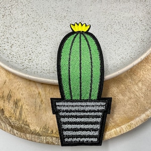 Peut inclure: Un cactus vert avec une fleur jaune sur le dessus, dans un pot à rayures noires et blanches. Le cactus est brodé sur un patch en tissu.