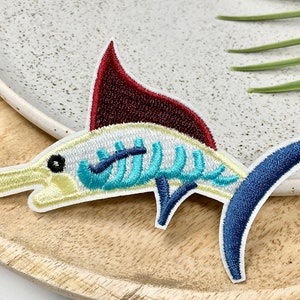 May include: A colorful embroidered iron-on patch of a blue and yellow marlin fish with a red dorsal fin. The patch is perfect for adding a nautical touch to clothing, bags, or other fabric items.