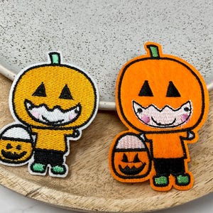 Peut inclure: Deux écussons brodés représentant des personnages de citrouilles d'Halloween. L'un est orange, l'autre jaune. Chacun tient un seau à bonbons. Les écussons ont des yeux et une bouche noirs, des pieds verts, sur un plateau en bois.