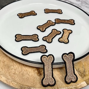 May include: Embroidered dog bone patches in gold with black borders, displayed on a white plate with a black rim. The patches are of varying sizes and are arranged on a wooden tray.