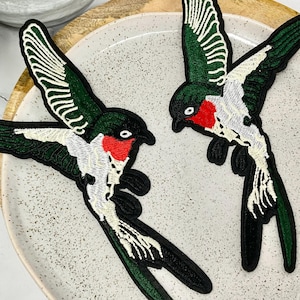 Peut inclure: Deux patchs brodés thermocollants représentant des oiseaux verts et blancs avec des accents rouges. Les oiseaux sont en position de vol, les ailes déployées.