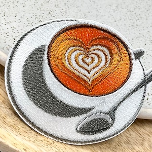 May include: A white embroidered patch of a coffee cup with a heart design in the coffee. The coffee is orange and the heart is white.