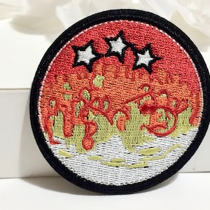 May include: A round embroidered patch with a black border. The center of the patch features a red and orange sunset with three white stars above it. The bottom of the patch is white with a yellow wavy line.