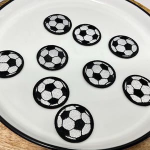 May include: A collection of nine black and white soccer ball patches arranged on a white plate. The patches are round with a classic soccer ball design, featuring black pentagons and white hexagons. The plate has a black rim and sits on a wooden surface.