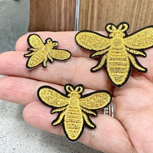 May include: Three gold embroidered bee patches of different sizes. The bees are outlined in black thread and have a textured, embroidered design.