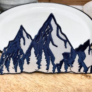May include: A blue embroidered patch depicting a mountain range with white snow-capped peaks. The patch is ideal for adding a touch of nature to clothing, bags, or other accessories.