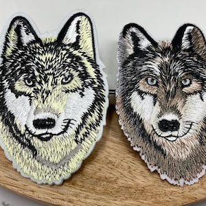 May include: Two embroidered patches featuring detailed wolf heads. The left patch displays a wolf with light yellow, white, and black thread. The right patch shows a wolf with brown, white, and black thread. Both have white borders.