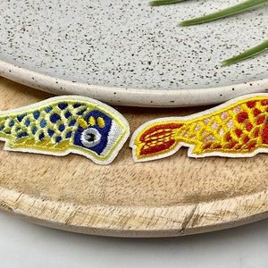 Peut inclure: Deux patchs brodés thermocollants en forme de poissons koï. Un poisson est jaune avec des détails bleus et blancs, et l'autre est rouge avec des détails blancs et jaunes.