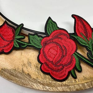 May include: Embroidered red rose patches with green leaves and black outlines. The patches are placed on a wooden plate with a white plate rim. The roses are in various stages of bloom.