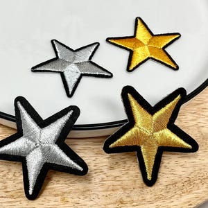 May include: Four embroidered iron-on patches in the shape of stars. Two stars are silver with black outlines and two stars are gold with black outlines.